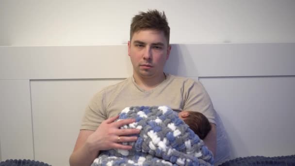 Виснажений молодий тато ковтає дитину на руках у ліжку. Чоловік з недбалим втомленим поглядом з новонародженим на руках . — стокове відео