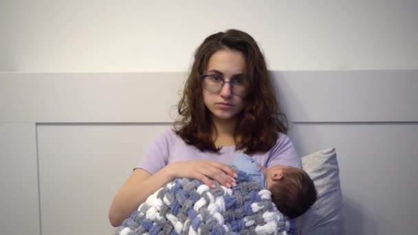 Egy kimerült fiatal anya a karjaiban ringat egy gyereket az ágyban. Egy őrült, fáradt tekintetű nő, egy újszülöttel a karjában.. — Stock videók