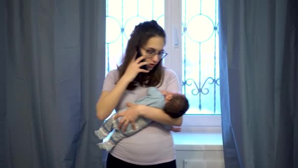 Une jeune mère secoue un enfant dans ses bras sur le fond d'une fenêtre. La femme ne sait pas quoi faire et parle au téléphone sur ses nerfs. — Video