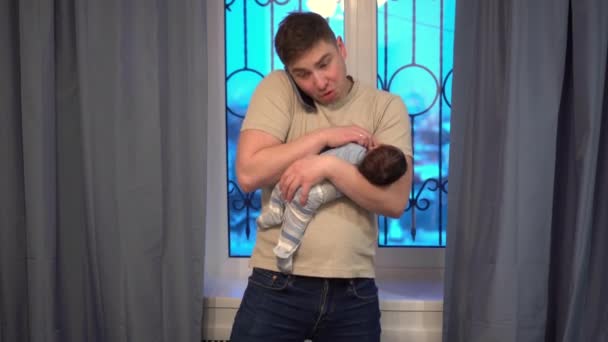 Un jeune père berçant un enfant dans ses bras sur le fond de la fenêtre. L'homme ne sait pas quoi faire et parle au téléphone sur ses nerfs. — Video