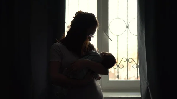 A young mother holds a child in her arms against the background of a window. The light from the window illuminates the image of a woman with a newborn baby. — Stock Photo, Image