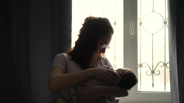 A young mother holds a child in her arms against the background of a window. The light from the window illuminates the image of a woman with a newborn baby. — Stock Photo, Image