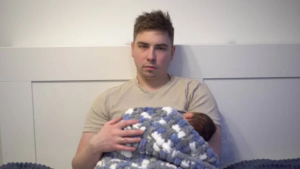 Un padre joven agotado acuna a un niño en sus brazos en la cama. Un hombre con una mirada frenética y cansada con un recién nacido en sus brazos. — Foto de Stock