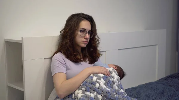 Una madre joven y agotada acuna a un niño en sus brazos en la cama. Una mujer con una mirada frenética y cansada con un recién nacido en sus brazos. — Foto de Stock