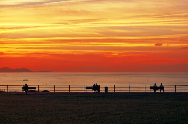 People relaxing on benches viewing the sea at sunset — Stock Photo, Image