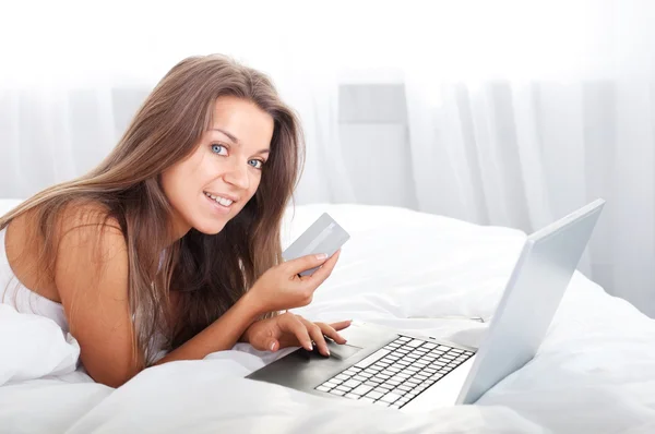 Молодая женщина, использующая ноутбук в постели с кредитной машиной — стоковое фото
