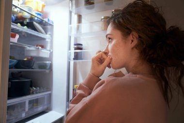 Undecided young caucasian woman checking fridge for some food at night clipart