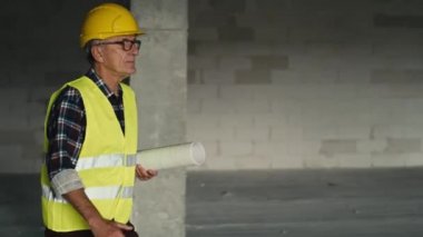 Kafkasyalı kıdemli mühendis inşaat alanında yürüyor ve planları tutuyor. 8K 'da kırmızı helyum kamerayla çekildi.     
