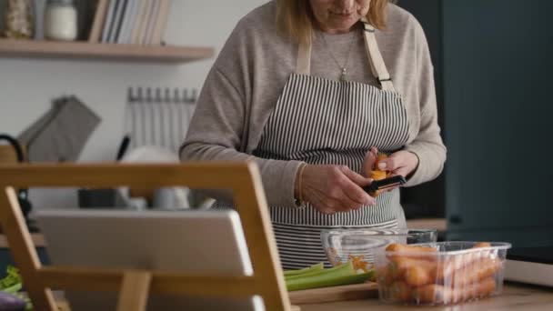 キッチンで料理をしながらニンジンを剥離白人のシニア女性 8Kでレッドヘリウムカメラで撮影 — ストック動画