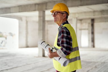 Kafkasyalı kıdemli mühendis inşaat sahasında yürüyor ve planları tutuyor