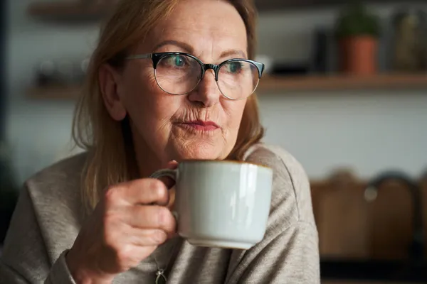 キッチンに座ってコーヒーを飲みながら目を離す思慮深い白人のシニア女性のクローズアップ — ストック写真