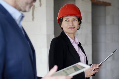 Beyaz kadın mühendis ve iş kadını inşaat sahasında dururken ellerinde belge tutuyorlar.