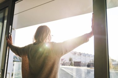 Back view of senior woman opening balcony doors in the beams of the sun