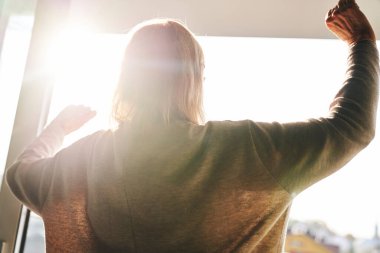 Back view of senior woman opening balcony doors in the beams of the sun