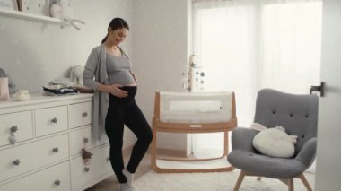 Hamileliği ilerlemiş beyaz bir kadın bebeğin odasında beşiğin yanında duruyor. 8K 'da kırmızı helyum kamerayla çekildi..