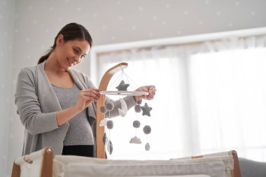 Beyaz hamile kadın bebeğin beşiğinin üzerine oyuncak yerleştiriyor.