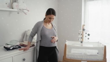 Gelişmiş hamilelik dönemindeki düşünceli beyaz kadın beşiğin yanında, bebek odasında duruyor. 8K 'da kırmızı helyum kamerayla çekildi..