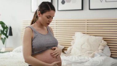 Hamileliği ilerlemiş beyaz bir kadın yatakta oturuyor ve güçlü krampları ve sırt ağrıları var. 8K 'da kırmızı helyum kamerayla çekildi.. 