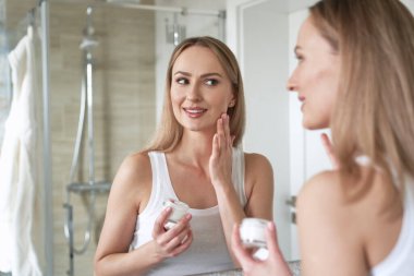 Genç beyaz kadın gülümsüyor ve aynadaki yansımasına yüz kremi sürüyor.