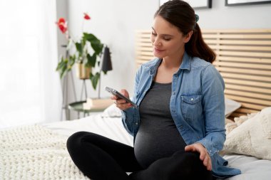 Yatakta cep telefonu kullanan beyaz bir kadın hamile.