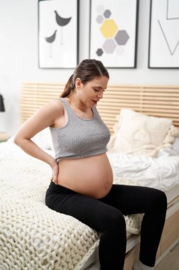 Hamileliği ilerlemiş beyaz kadın yatakta oturuyor ve güçlü krampları ve sırt ağrısı var. 