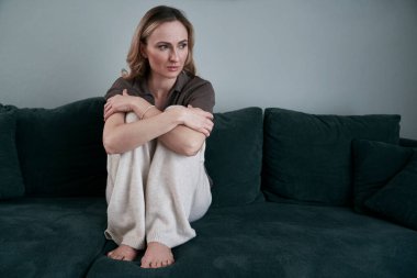 Evde depresyondaki bir kadın kanepeye çömelmiş ve aşağıya bakıyor.