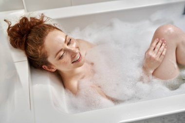 Yüksek açılı genç kızıl saçlı bir kadın banyo yapıyor ve başka tarafa bakıyor.