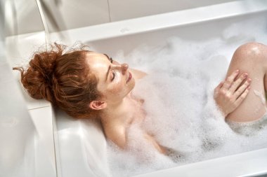 Kızıl saçlı kadın gözleri kapalı banyo yapıyor. 