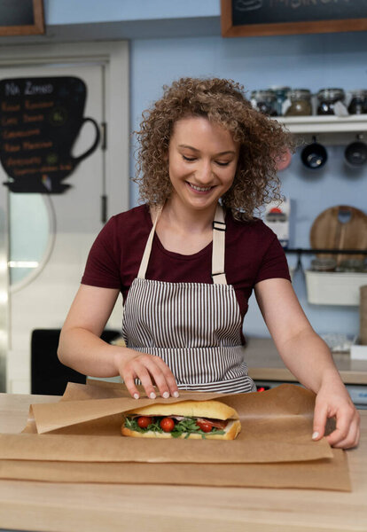 Young caucasian waitress packing prepared sandwich in commercial kitchen