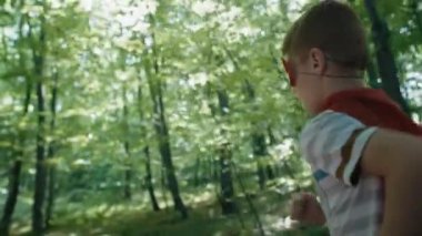 Ormanda koşan süper kahraman kostümü giyen beyaz bir çocuk. 4K 'da kırmızı helyum kamerayla çekildi..     