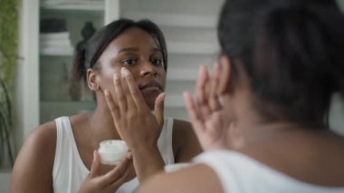 Young African-American  woman applying face cream in the mirror reflection. Shot with RED helium camera in 8K.  