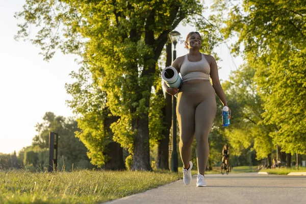 stock image Young African American woman walking with exercise mat and bottle of water through the park in a summer day