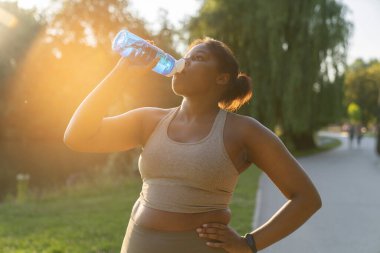Thirsty African American woman drinking water during the workout at the park 