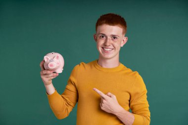 Red head caucasian teenage boy pointing at piggy bank