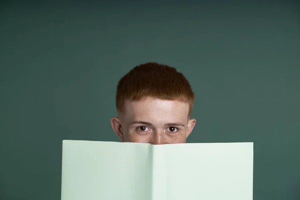 Close up of caucasian male teenager hidden behind green paper notebook