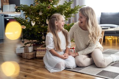 Caucasian girl and mother having fun while sitting next to Christmas tree