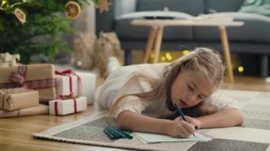Noel ağacının yanında halıya uzanmış beyaz bir kız Noel Baba 'ya mektup yazıyor. 8K 'da kırmızı helyum kamerayla çekildi.. 