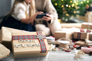 Noel hediyesinin ayrıntıları Noel ağacının altında yatıyor ve anne ile kızı da arka planda. 