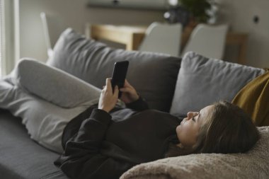 Kafkasyalı kadın kanepeye uzanıp cep telefonu kullanıyor.