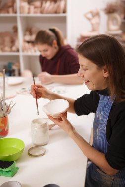 Çömlekçilik dersinde seramik kâse boyarken gülen bir kadının dikey görüntüsü.