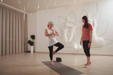 Ağaçta dikilen yaşlı kadın, yoga stüdyosunda kişisel antrenörle birlikte asana pozu veriyor.