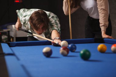 Genç adam bilardo topuna ıstakayla vurmaya hazırlanıyor, kız arkadaşıyla bilardo oynuyor.