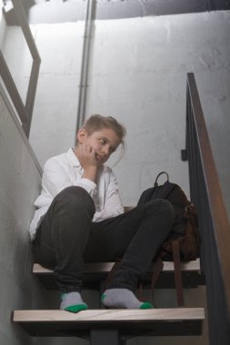 Üzgün görünen, merdivenlerde tek başına oturan genç bir çocuğun dikey uzun bir görüntüsü.