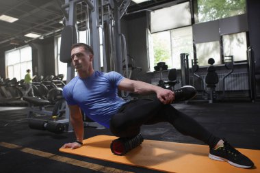 Professional bodybuilder using foam roller, stretching body muscles after workout at gym