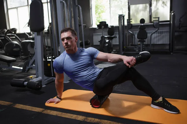 Focused male athlete using foam roller, stretching muscles after exercising at gym