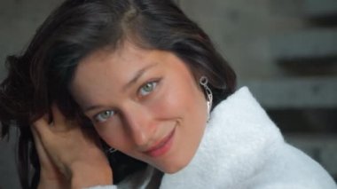 Genç ve güzel Avrupalı uzun saçlı esmer kız sonbahar elbiseleriyle merdivenlerde oturuyor ve beyaz bir süveter ve elleriyle kendine dokunuyor.