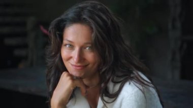 Genç ve güzel Avrupalı uzun saçlı esmer kız güz kıyafetleriyle bankta oturuyor ve beyaz bir süveter ve gülümsüyor.