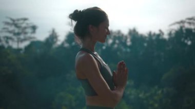 Çekici Avrupalı esmer spor kızı doğada yoga yapıyor. Havuzun kenarında yeşil bir yağmur ormanı var ve derin derin nefes alıyor..