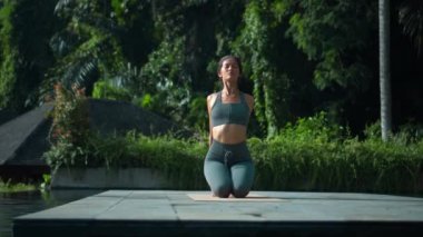 Çekici Avrupalı esmer spor kızı doğada yoga yapıyor. Havuzun kenarında yeşil bir yağmur ormanı var ve derin derin nefes alıyor..