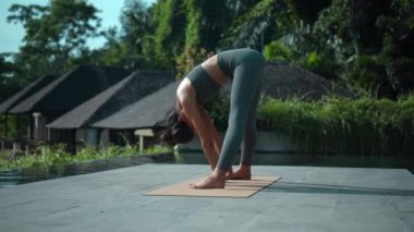 Çekici Avrupalı esmer spor kızı doğada yoga yapıyor. Havuzun kenarında yeşil bir yağmur ormanı var ve derin derin nefes alıyor..